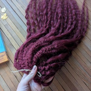 May include: A bundle of long, burgundy-colored synthetic hair extensions. The hair is curly and textured, and is tied together with a string.