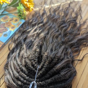 May include: Dark brown curly hair extensions with a sewn base and a white ribbon. The extensions are arranged on a wooden surface, with a blurred background featuring sunflowers and a painting of sunflowers.