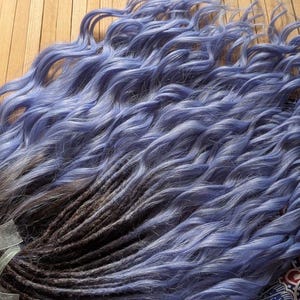May include: A bundle of blue and brown dreadlocks with wavy ends. The dreadlocks are a mix of blue and brown, with a gradient effect. The dreadlocks are lying on a wooden surface, with a colorful patterned fabric and a book with sunflowers in the background.