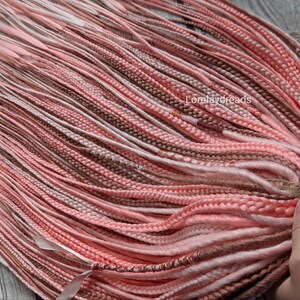 May include: A bundle of synthetic hair extensions in shades of pink, brown, and white. The hair is braided and ready to be added to natural hair.