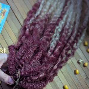May include: A bundle of curly hair extensions with a burgundy ombre to blonde color. The hair is tied together with a brown string.