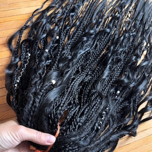Puede incluir: Un conjunto de extensiones de cabello sintético negro que presenta varios estilos de trenzado, incluyendo trenzas de caja y torzadas. Algunas trenzas están adornadas con pequeñas cuentas blancas y costuras decorativas. El cabello se sujeta con un hilo naranja.