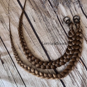 Puede incluir: Dos trenzas largas marrones con anillos metálicos en los extremos, dispuestas sobre una superficie de madera. Las trenzas miden aproximadamente 90 cm de largo.