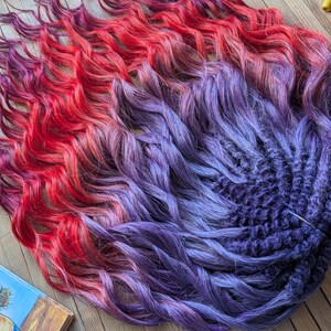May include: A bundle of vibrant, wavy hair extensions in a gradient of colors. The colors transition from red to purple to deep blue. The extensions are styled in a loose, flowing pattern, with a textured appearance.