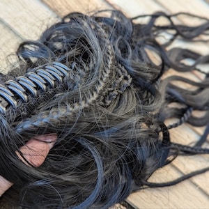 Puede incluir: Extension de pelo sintético negro con un clip de sujeción. El pelo es largo y ondulado, y el clip es de plástico.