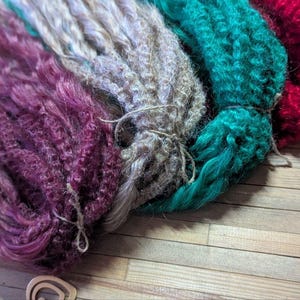 May include: Four bundles of synthetic hair extensions in various colors, including purple, beige, teal, and red. The hair is tied together with string.