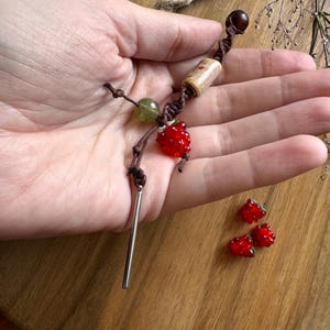 Peut inclure: Un collier fait à la main avec un pendentif fraise en verre rouge, une perle verte et une perle en bois. Le collier est sur un cordon marron avec une barre argentée. Trois perles de fraise supplémentaires sont également visibles.