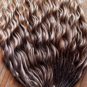 May include: A bundle of wavy, ombre hair extensions. The hair transitions from light blonde at the top to shades of brown and dark brown at the bottom. The extensions are tied together with a white string and rest on a wooden surface.