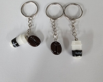 Könnte beinhalten: Drei Metallschlüsselanhänger mit Miniatur-Kaffeetassen in Weiß mit schwarzer Banderole und der Aufschrift Coffee sowie dunkelbraunen Kaffeebohnen-Anhängern. Jeder Schlüsselanhänger besteht aus einem silbernen Ring an einer kurzen Kette.