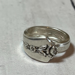 May include: Silver-toned ring with a floral design. The ring appears to be crafted from a spoon, with the spoon's handle forming the band and the bowl of the spoon featuring a raised floral pattern. The ring has a vintage or antique aesthetic.