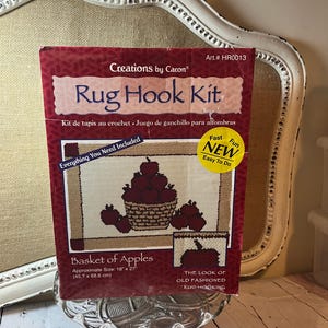 May include: A rug hook kit by Creations by Caron, featuring a "Basket of Apples" design. The kit includes everything needed and is approximately 18" x 27" in size. The packaging is red and features a picture of the finished rug.