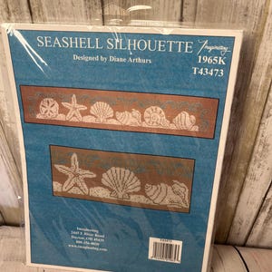 May include: A cross-stitch pattern titled "Seashell Silhouette" by Diane Arthurs. The design features a brown border with white seashell and starfish motifs on a blue background. The package is sealed in clear plastic.