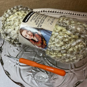 May include: A skein of variegated green, yellow, and white chunky yarn with a paper label reading Country Loom Colorburst, Yarn Bee, and 47 yds/3.5 oz, paired with an orange crochet hook marked H-5.00mm, resting inside a clear glass bowl.