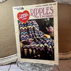 May include: A vintage crochet pattern book titled "Ripples By Design" featuring a colorful afghan design. The cover displays a completed blanket in shades of blue, purple, and yellow. The book is on a glass cake stand.