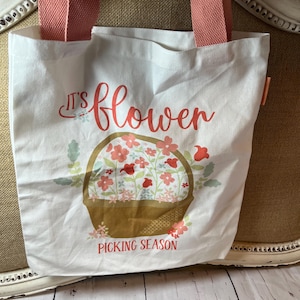 May include: White canvas tote bag with coral pink handles. The bag features a floral design with a basket of flowers and the words "It's flower" and "Picking Season" in a script font. The bag is on a neutral background.