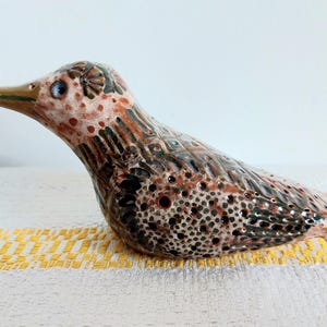 Puede incluir: Figura de pájaro de cerámica con un patrón moteado en tonos marrón, negro y crema. El pájaro tiene un pico largo y curvo y un ojo pequeño y redondo. La figura está sobre una superficie a rayas blancas y amarillas.