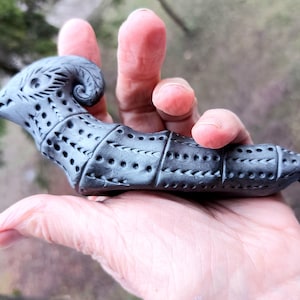 Könnte beinhalten: Eine handgeschnitzte, graue Skulptur eines stilisierten Vogels oder Fabelwesens. Die Skulptur weist komplizierte Ausschnittmuster und ein eingerolltes Horn auf. Das Objekt wird in einer Hand gehalten und zeigt seine detaillierte Handwerkskunst und sein einzigartiges Design.