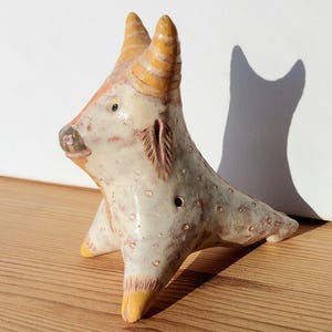 May include: A ceramic bull figurine with a cream-colored body, speckled with brown spots. The horns and hooves are yellow, and the face has pink and orange accents. The sculpture has a unique, handcrafted appearance.