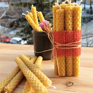 Puede incluir: Un manojo de velas de cera de abeja natural con un patrón de panal, atado con una cinta de arpillera roja. Las velas están dispuestas en un jarrón y sobre una mesa de madera.