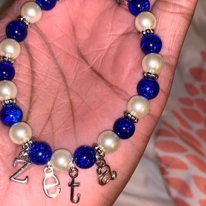 Puede incluir: Un brazalete de cuentas azules y blancas con dijes plateados que deletrean "Zeta".