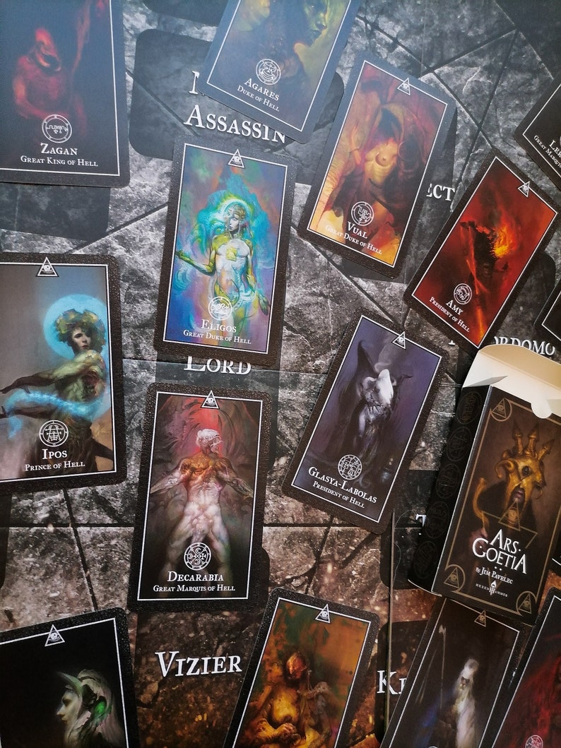 May include: A set of tarot cards featuring illustrations of demons and their titles. The cards are black with gold accents and feature a variety of demon imagery, including a demon with horns and a crown, a demon with wings and a sword, and a demon with a skull and crossbones. The cards are arranged in a random order on a dark surface.