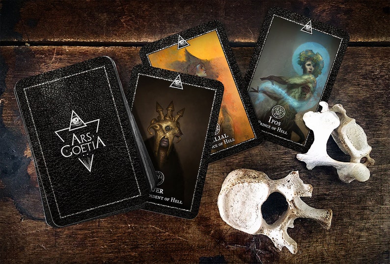 May include: A black leather-bound box with the text "ARS GOETIA" on the front.  There are four cards on a wooden surface, each with a different image and text.  The cards are from a deck of tarot cards.  The cards are: "LILIAL, Duke of Hell", "IPOS, Prince of Hell", "BUER, President of Hell", and "Astarte, Countess of Hell".