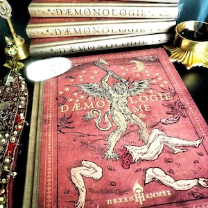 Puede incluir: Una pila de cuatro libros con lomos dorados titulados DÆMONOLOGIE, coronada por un libro de tapa dura rojo con una ilustración de un demonio alado, el texto DÆMONOLOGIE INFORME y la palabra HEXENHAMMER, junto a un quemador de incienso dorado.