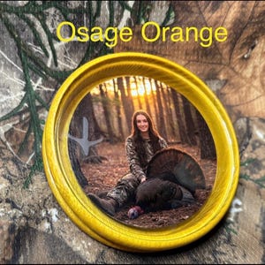 May include: A yellow circular frame with the words "Osage Orange" in yellow text. Inside the frame is a woman in camouflage clothing sitting next to a turkey. The background shows a forest.