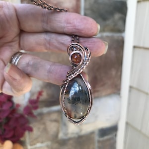 Puede incluir: Un colgante de alambre trenzado que presenta una piedra de ágata dendrítica ovalada y lisa con inclusiones oscuras en forma de árbol y una cuenta redonda más pequeña de color ámbar translúcido en la parte superior, todo en cobre pulido.