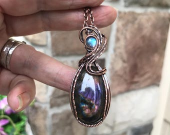 Puede incluir: Un colgante con una gran piedra preciosa de labradorita ovalada con destellos iridiscentes púrpuras y azules, envuelta en intrincado alambre de cobre. Un cabujón de labradorita más pequeño y redondo se sitúa sobre la piedra principal, también en cobre.