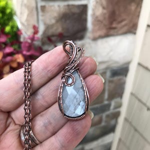 May include: A handcrafted copper wire-wrapped pendant necklace featuring a faceted, teardrop-shaped, gray gemstone. The pendant is suspended from a matching copper chain. The necklace is held in a hand.
