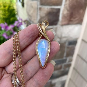 May include: A teardrop-shaped moonstone pendant featuring a blue iridescent sheen, encased in a gold-toned wire-wrapped setting with a small copper accent at the base, suspended from a matching gold-toned metal link chain necklace.