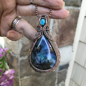 Peut inclure: Un pendentif en labradorite en forme de goutte enveloppé dans un fil de cuivre complexe. Le pendentif présente une grande pierre irisée bleue et grise à la base et un cabochon de labradorite bleue ovale plus petit fixé au sommet, suspendu à une chaîne.