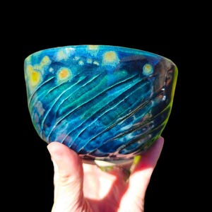 Puede incluir: Un cuenco de cerámica hecho a mano con un diseño texturizado y acanalado. El cuenco presenta una vibrante paleta de colores azules y verdes, con acentos amarillos. El cuenco se sostiene en una mano sobre un fondo negro.