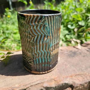 May include: A handmade ceramic tumbler with a textured, ridged surface. The glaze features a blend of teal, brown, and black colors. The cup is cylindrical and appears to be suitable for beverages. The tumbler is sitting on a stone surface.