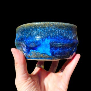 May include: A handmade ceramic bowl with a vibrant blue glaze. The bowl's exterior features a textured, layered effect with shades of deep and light blue, and a touch of gold. The bowl is held in a hand against a black background.