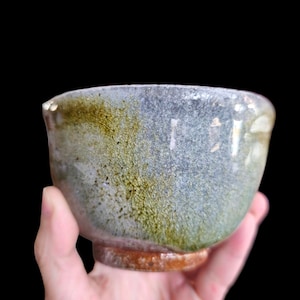 Può includere: Una ciotola in ceramica fatta a mano con una smaltatura unica. La ciotola presenta una miscela di tonalità blu, verdi e marroni, con una superficie strutturata. La ciotola è tenuta in mano su uno sfondo nero. La ciotola viene probabilmente utilizzata per servire cibo o come oggetto decorativo.