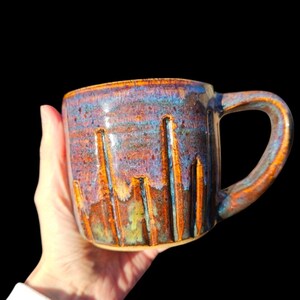 Puede incluir: Una taza de cerámica hecha a mano con un esmalte único. La taza presenta una superficie texturizada con ranuras verticales y un asa. El esmalte combina colores azules, naranjas y morados, creando un diseño atractivo. La taza se sostiene sobre un fondo negro.