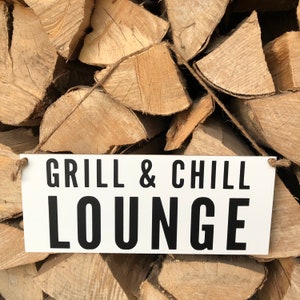 Könnte beinhalten: Ein weißes Holzschild mit schwarzem Text, auf dem "Grill & Chill Lounge" steht, hängt an einem Stapel Brennholz.