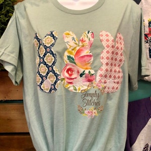 May include: A light green t-shirt with three bunny-shaped fabric appliques. The bunnies are made of different patterned fabrics, including floral, geometric, and plaid. The shirt also features a small, circular logo that says "Simply Stitches" with a floral wreath around it.
