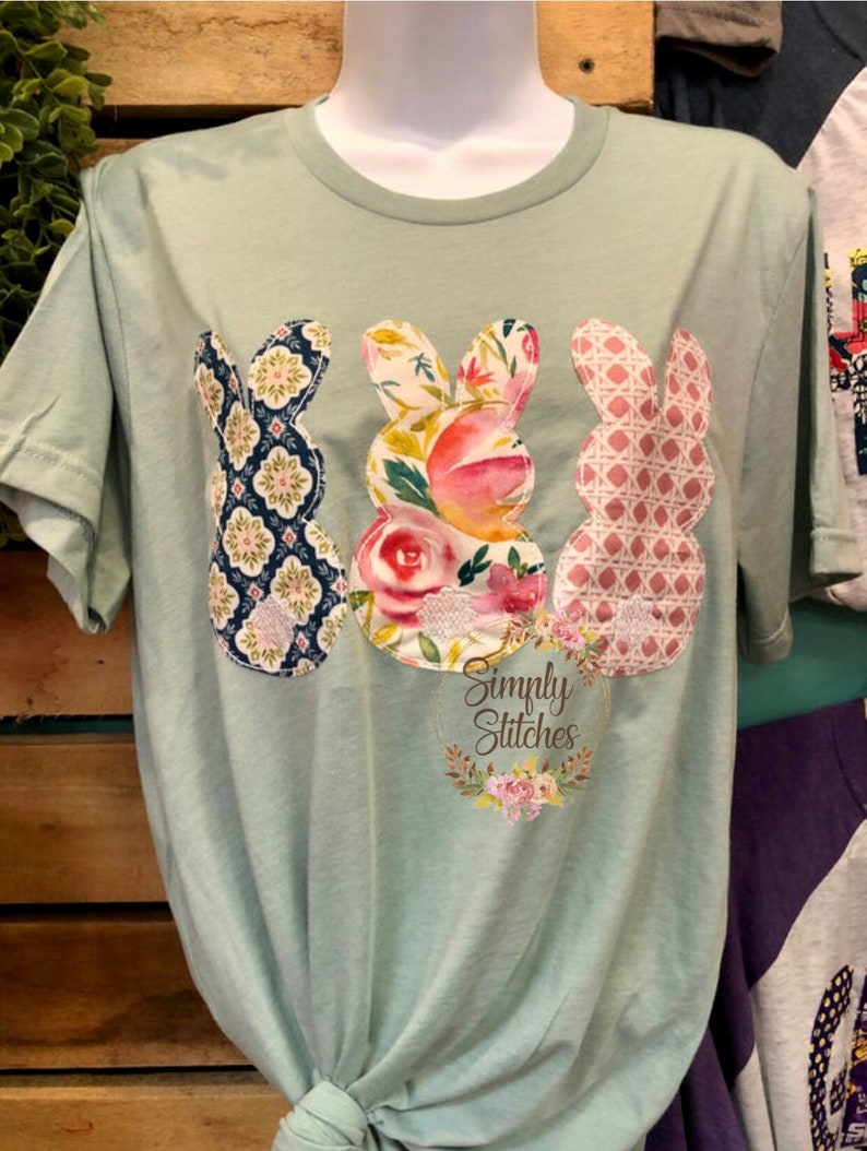 May include: A light green t-shirt with three bunny-shaped fabric appliques. The bunnies are made of different patterned fabrics, including floral, geometric, and plaid. The shirt also features a small, circular logo that says "Simply Stitches" with a floral wreath around it.