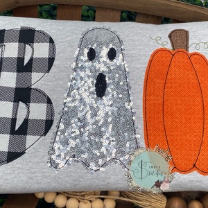 Puede incluir: Una almohada gris con la palabra "BOO" bordada. La "B" es de cuadros negros y blancos, la "O" es un fantasma blanco con lentejuelas plateadas y la "O" es una calabaza naranja.