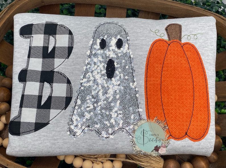 May include: A gray pillow with the word "BOO" embroidered on it. The "B" is black and white checkered, the "O" is a white ghost with silver sequins, and the "O" is an orange pumpkin.