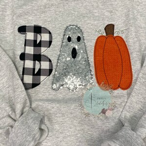 May include: A gray sweatshirt with a black and white checkered "B", a silver sequined ghost, and an orange pumpkin with a green stem that says "BOO" on it.