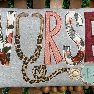 May include: A gray sweatshirt with the word "NURSE" spelled out in colorful fabric letters. A stethoscope is sewn onto the sweatshirt, with a leopard print design.