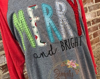 May include: A gray and red baseball tee shirt with the words "Merry and Bright" in colorful fabric letters. The letters are outlined in black thread. The shirt has a floral design with the words "Simply Stitches" in the bottom right corner.