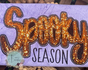 May include: Purple t-shirt with the words "Spooky Season" in black text. The word "Spooky" is made of orange sequins.
