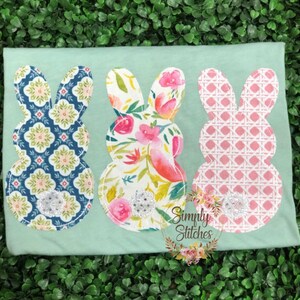 May include: Three pastel Easter bunny shapes sewn onto a light green t-shirt. The bunnies are made of different patterned fabrics: blue and white geometric, floral, and pink and white checkered. Each bunny has a silver glitter button in the center.