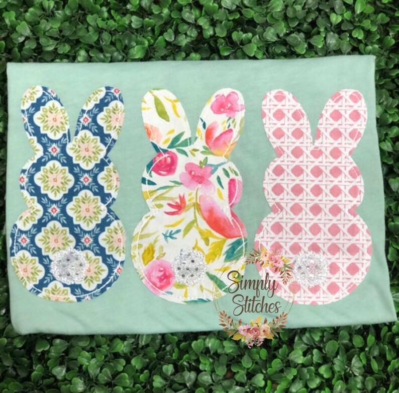 May include: Three pastel Easter bunny shapes sewn onto a light green t-shirt. The bunnies are made of different patterned fabrics: blue and white geometric, floral, and pink and white checkered. Each bunny has a silver glitter button in the center.