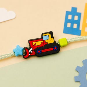 May include: A light green woven cord bracelet featuring a central rubber charm of a red and yellow bulldozer, flanked by a small blue star-shaped bead on the left and a lime green cube-shaped bead on the right, secured on a thin cord.